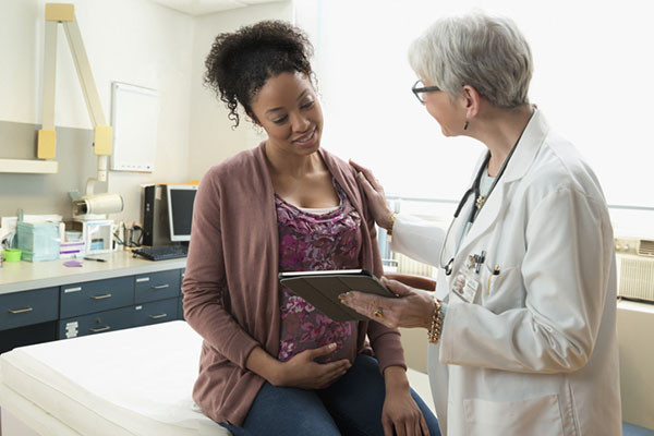 photo of a pregnant person in an examination room speaking with a gynecologist, who is holding a tablet and showing it to the patient